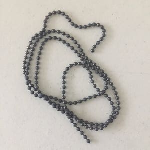 May include: A long chain of small, dark gray metal beads. The chain is coiled and laid out on a white surface.