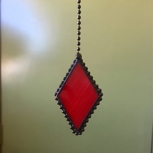 May include: A red stained glass diamond hanging from a chain. The diamond is framed with a black metal border.