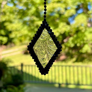 May include: A diamond-shaped stained glass suncatcher with a black beaded border and a textured, yellow-green center. The suncatcher hangs from a black beaded chain, with a blurred green background.