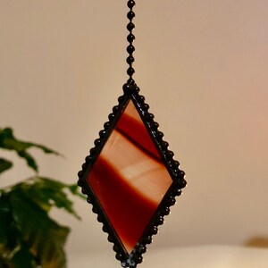 May include: A stained glass diamond-shaped suncatcher with a dark brown beaded border and a chain for hanging. The glass has a gradient of red and white hues. Decorative item for windows.