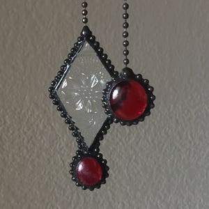 May include: A stained glass suncatcher with a diamond-shaped center piece and two round red glass accents. The suncatcher is suspended from a black chain.