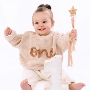Peut inclure: Une petite fille portant un pull en tricot beige avec le mot "un" brod&eacute; dessus, assise devant un grand chiffre un en bois blanc. Elle tient une baguette rose et blanche avec un sommet en forme d'&eacute;toile qui dit "Joyeux anniversaire".