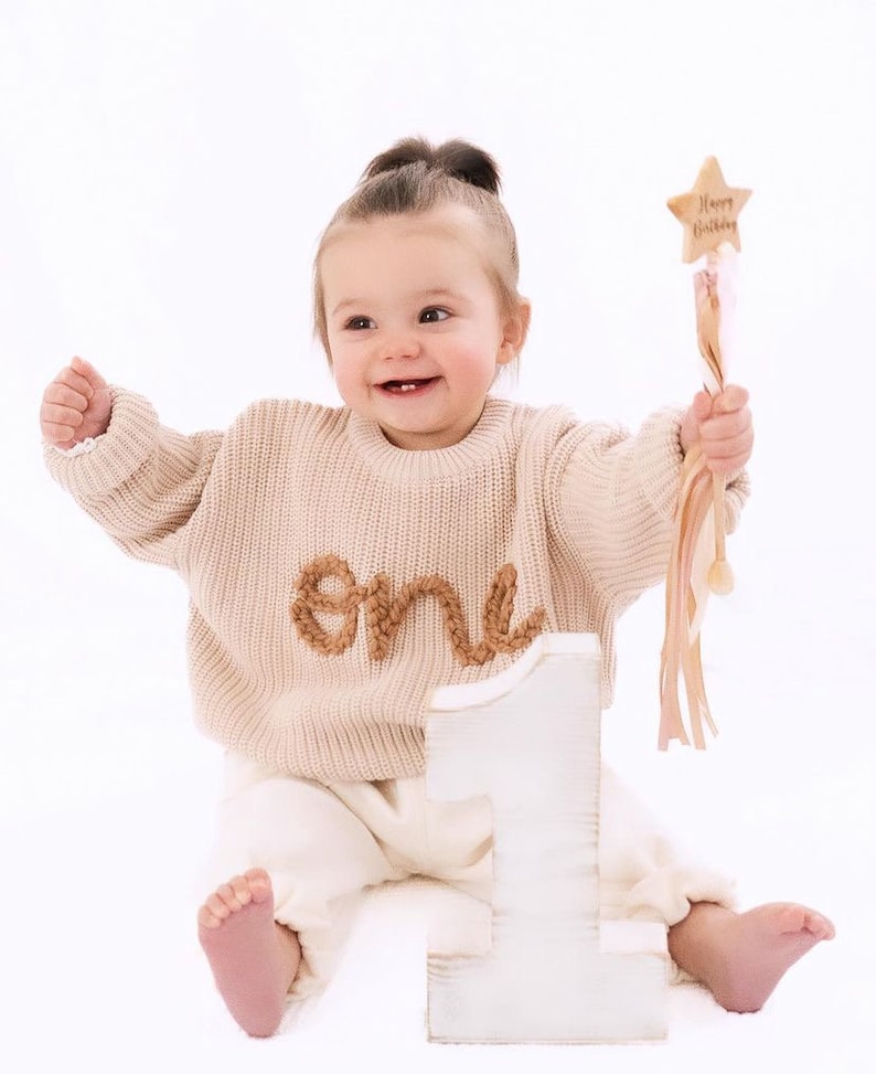 Peut inclure: Une petite fille portant un pull en tricot beige avec le mot "un" brod&eacute; dessus, assise devant un grand chiffre un en bois blanc. Elle tient une baguette rose et blanche avec un sommet en forme d'&eacute;toile qui dit "Joyeux anniversaire".