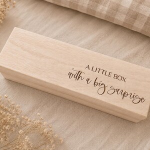 Könnte beinhalten: Eine rechteckige, helle Holzschachtel mit Klappdeckel. Auf der Oberseite befindet sich der eingravierte Text A LITTLE BOX with a big surprise in einer Kombination aus Block- und Schreibschrift. Die Holzmaserung ist deutlich sichtbar.
