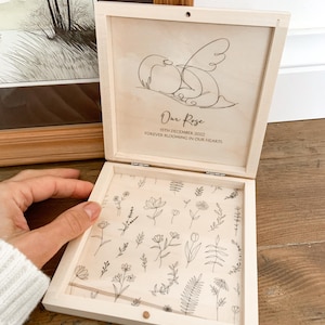 Peut inclure: Bo&icirc;te en bois clair ouverte, orn&eacute;e d'un dessin au trait d'oiseaux et de l'inscription "Our Rose". L'int&eacute;rieur est d&eacute;cor&eacute; de motifs floraux. La bo&icirc;te est tenue ouverte par une main.