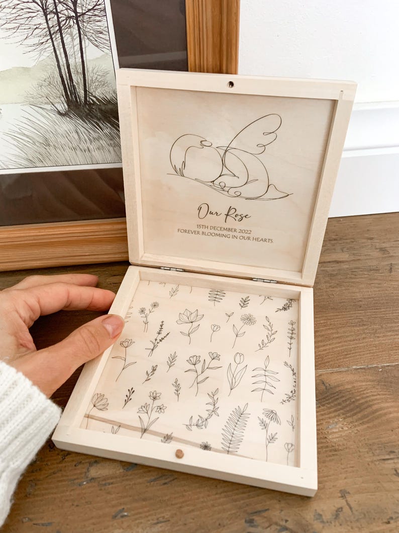 Peut inclure: Bo&icirc;te en bois clair ouverte, orn&eacute;e d'un dessin au trait d'oiseaux et de l'inscription "Our Rose". L'int&eacute;rieur est d&eacute;cor&eacute; de motifs floraux. La bo&icirc;te est tenue ouverte par une main.