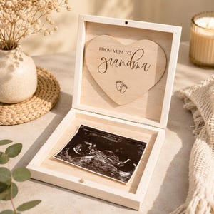 Könnte beinhalten: Eine helle Holzkiste mit einem herzförmigen Deckeleinsatz mit der Aufschrift "FROM MUM TO grandma" mit Babyfüßen. In der Kiste befindet sich ein schwarz-weißes Ultraschallbild. Eine Kerze und eine Vase mit Blumen sind im Hintergrund.