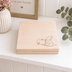 Peut inclure: Une boîte en bois de couleur claire avec un simple dessin au trait d'un ange endormi sur le dessus. La boîte est carrée et présente un fini grain de bois naturel. La boîte est posée sur une surface blanche.