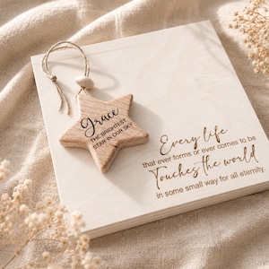 Könnte beinhalten: Eine quadratische Platte aus hellem Holz mit dem eingravierten Text 'Every life that ever forms or ever comes to be Touches the world in some small way for all eternity.' Ein hölzerner Stern mit dem Text 'Grace THE BRIGHTEST STAR IN OUR SKY' hängt an einer Juteschnur.