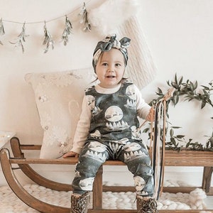 Peut inclure: Un b&eacute;b&eacute; portant une combinaison bleu fonc&eacute; et blanche avec un imprim&eacute; de lune et de for&ecirc;t. La combinaison est port&eacute;e sur un t-shirt blanc &agrave; manches longues. Le b&eacute;b&eacute; est assis sur un tra&icirc;neau en bois et porte un bandeau bleu fonc&eacute; avec un n&oelig;ud. Le b&eacute;b&eacute; tient un b&acirc;ton en bois avec des rubans color&eacute;s.