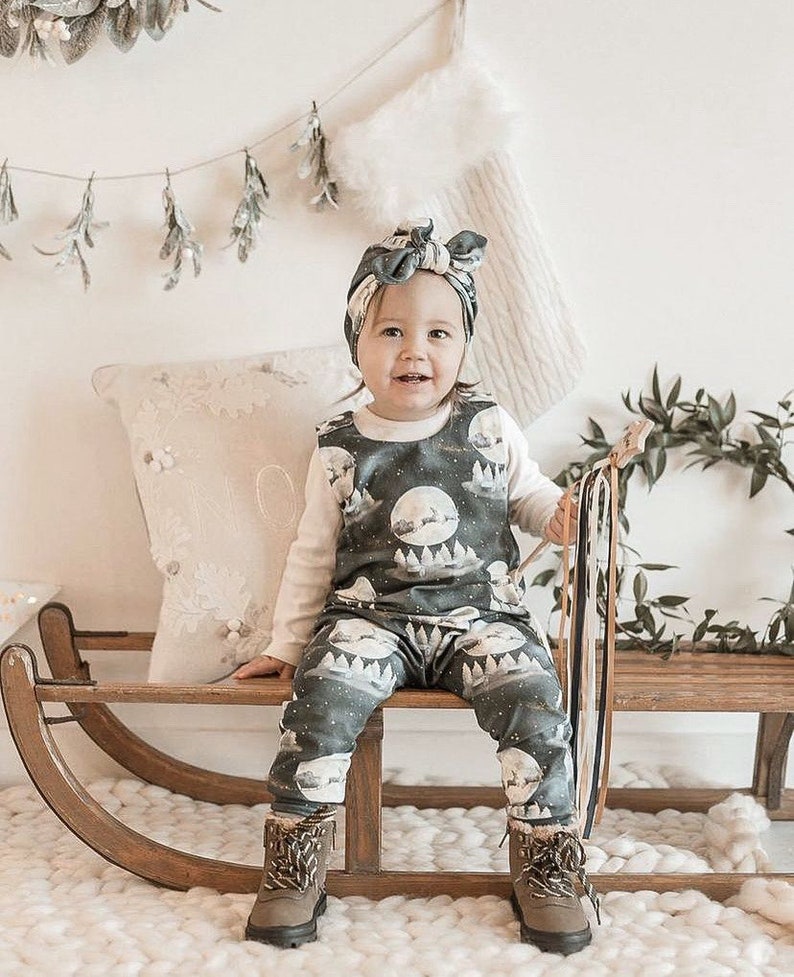 Peut inclure: Un b&eacute;b&eacute; portant une combinaison bleu fonc&eacute; et blanche avec un imprim&eacute; de lune et de for&ecirc;t. La combinaison est port&eacute;e sur un t-shirt blanc &agrave; manches longues. Le b&eacute;b&eacute; est assis sur un tra&icirc;neau en bois et porte un bandeau bleu fonc&eacute; avec un n&oelig;ud. Le b&eacute;b&eacute; tient un b&acirc;ton en bois avec des rubans color&eacute;s.