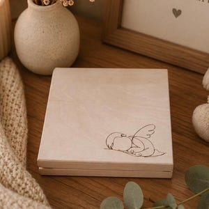 Peut inclure: Une bo&icirc;te en bois de couleur claire avec un couvercle &agrave; charni&egrave;re. La bo&icirc;te pr&eacute;sente un simple dessin au trait d'un ange endormi sur le devant. La bo&icirc;te est pos&eacute;e sur une surface en bois.