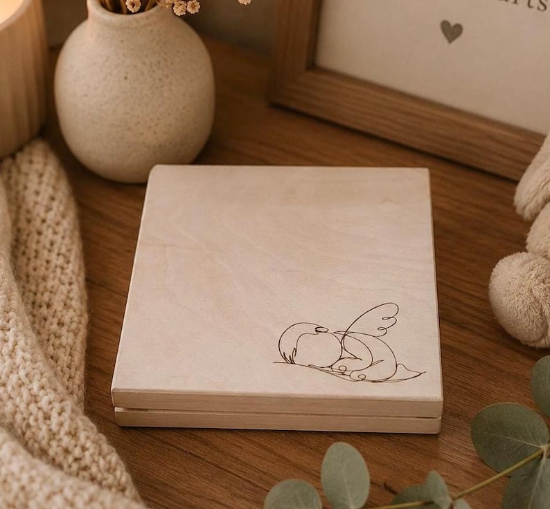 Peut inclure: Une bo&icirc;te en bois de couleur claire avec un couvercle &agrave; charni&egrave;re. La bo&icirc;te pr&eacute;sente un simple dessin au trait d'un ange endormi sur le devant. La bo&icirc;te est pos&eacute;e sur une surface en bois.