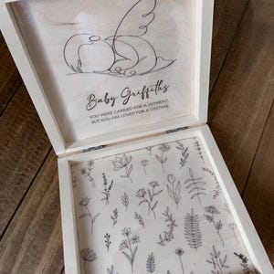 Peut inclure: Un coffret souvenir en bois de couleur claire avec un couvercle &agrave; charni&egrave;re. L'int&eacute;rieur du couvercle pr&eacute;sente un dessin au trait d'un ange et le nom "Baby Griffiths" avec le texte "YOU WERE CARRIED FOR A MOMENT, BUT YOU ARE LOVED FOR A LIFETIME." L'int&eacute;rieur de la bo&icirc;te est d&eacute;cor&eacute; de dessins au trait floraux.