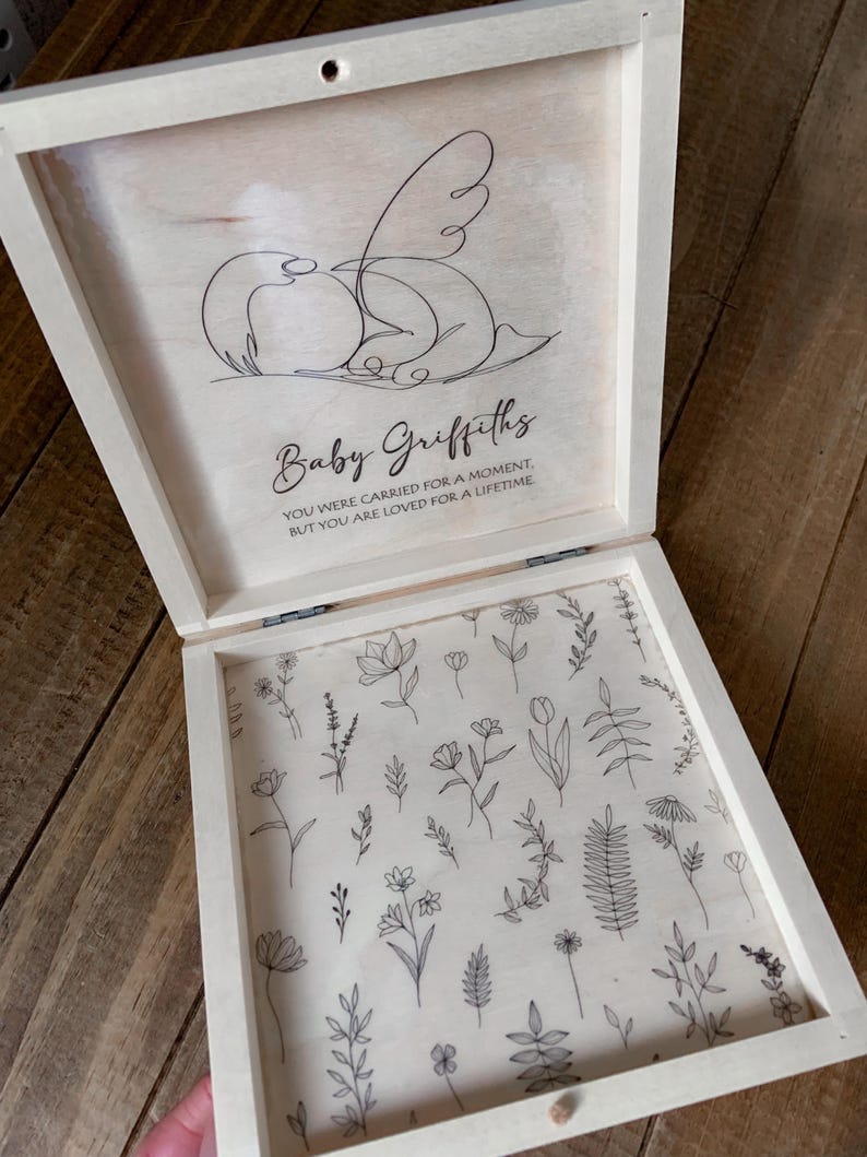 Peut inclure: Un coffret souvenir en bois de couleur claire avec un couvercle &agrave; charni&egrave;re. L'int&eacute;rieur du couvercle pr&eacute;sente un dessin au trait d'un ange et le nom "Baby Griffiths" avec le texte "YOU WERE CARRIED FOR A MOMENT, BUT YOU ARE LOVED FOR A LIFETIME." L'int&eacute;rieur de la bo&icirc;te est d&eacute;cor&eacute; de dessins au trait floraux.