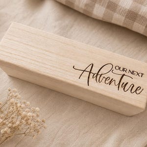 Puede incluir: Un bloque rectangular de madera de color claro con un acabado liso. La cara frontal presenta el texto grabado con láser "our next Adventure" en una combinación de una fuente sans-serif limpia y una tipografía cursiva decorativa y fluida.