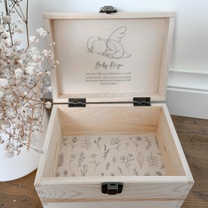 May include: A light-colored wooden keepsake box with a hinged lid, open to reveal a floral patterned interior. The lid features a drawing of a baby with wings and the name "Baby Reiga". The box is secured with a metal clasp.