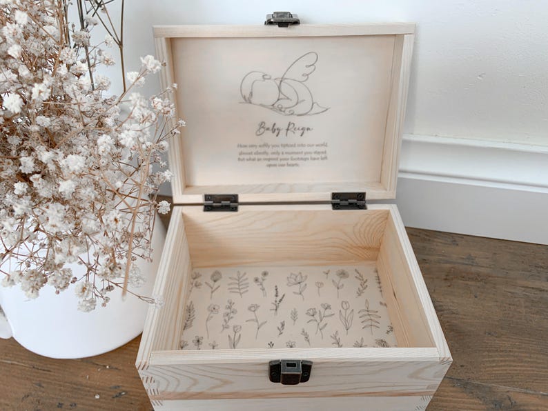 May include: A light-colored wooden keepsake box with a hinged lid, open to reveal a floral patterned interior. The lid features a drawing of a baby with wings and the name "Baby Reiga". The box is secured with a metal clasp.