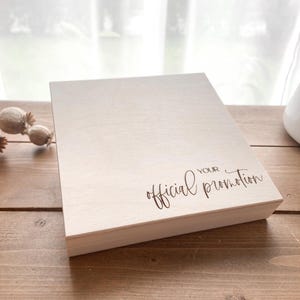 Könnte beinhalten: Eine hellfarbene Holzkiste mit den Worten "your official promotion" in eleganter Schrift. Die Kiste ist rechteckig und scheint aus unbehandeltem Holz zu sein. Die Kiste steht auf einer Holzoberfläche.