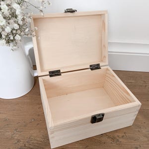 May include: An open, light-colored wooden box with a hinged lid and metal clasp. The box is empty and sits on a wooden surface. A white pitcher with white flowers is in the background.