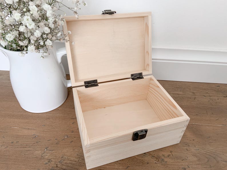May include: An open, light-colored wooden box with a hinged lid and metal clasp. The box is empty and sits on a wooden surface. A white pitcher with white flowers is in the background.