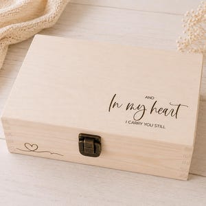 May include: A light-colored wooden box with a hinged lid and a metal clasp. The box is engraved with the words "In my heart and I carry you still" in a script font. A small heart and line design is on the lower left corner.