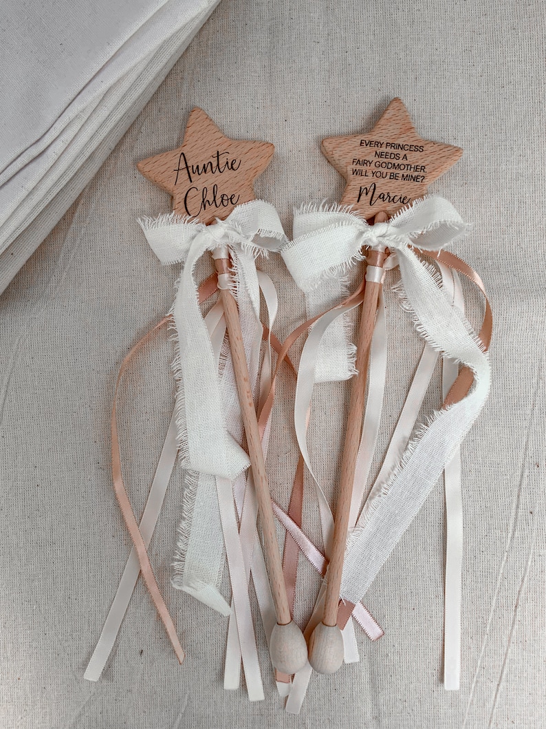 Puede incluir: Dos varitas de madera en forma de estrella con mangos de madera clara. Una est&aacute; grabada con "Auntie Chloe" y la otra con "EVERY PRINCESS NEEDS A FAIRY GODMOTHER. WILL YOU BE MINE?" y "Marcie". Ambas tienen lazos de cinta blanca deshilachada y cintas largas.