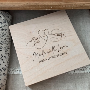Puede incluir: Una caja de madera de color claro con un diseño de corazón y jeringa, y las palabras "Made with Love, AND A LITTLE SCIENCE" grabadas en la superficie. La caja está dentro de un cajón de madera, junto a una tela con estampado floral.