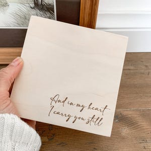May include: A wooden sign with the text "And in my heart I carry you still" engraved in brown lettering.