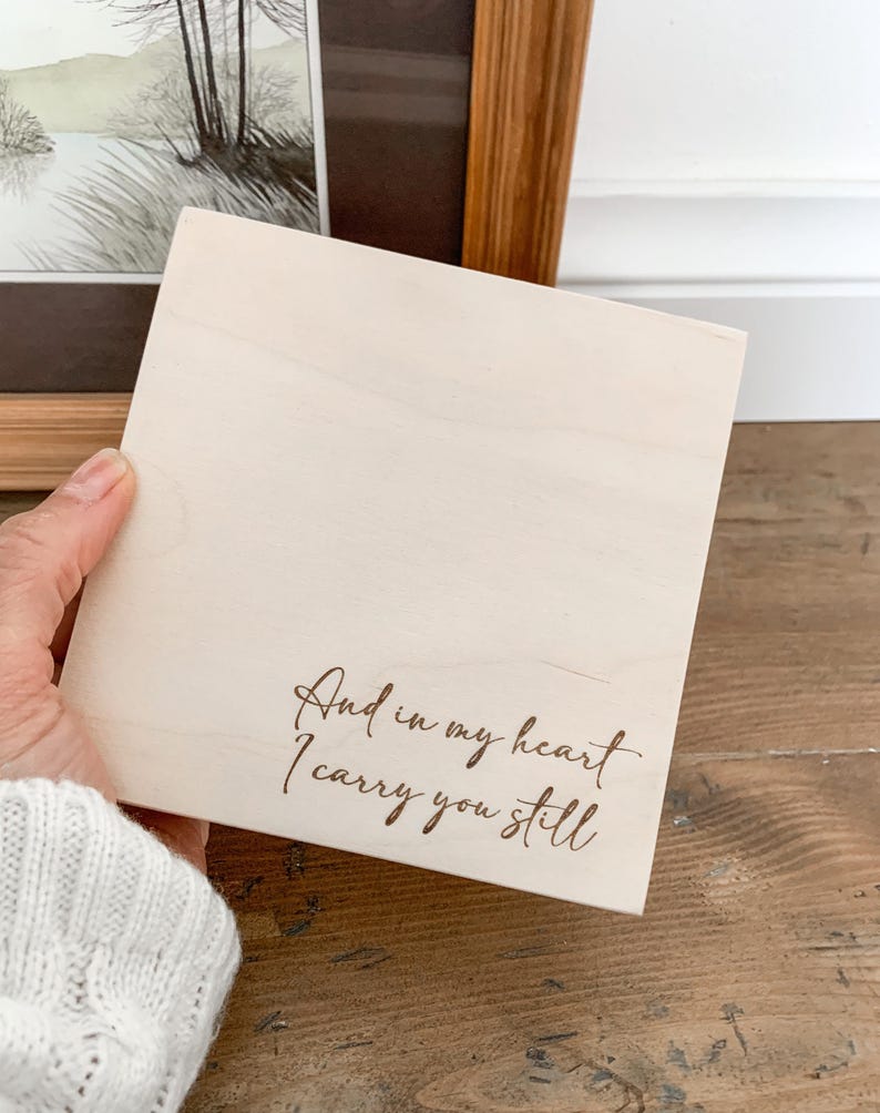 May include: A wooden sign with the text "And in my heart I carry you still" engraved in brown lettering.