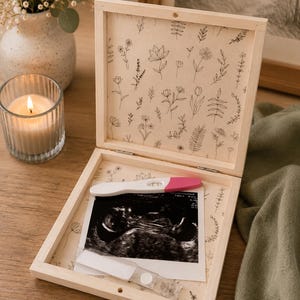 Peut inclure: Coffret souvenir en bois clair, orn&eacute; d'un motif floral &agrave; l'int&eacute;rieur. Il contient un test de grossesse et une &eacute;chographie. Une bougie allum&eacute;e et un vase de fleurs sont en arri&egrave;re-plan.