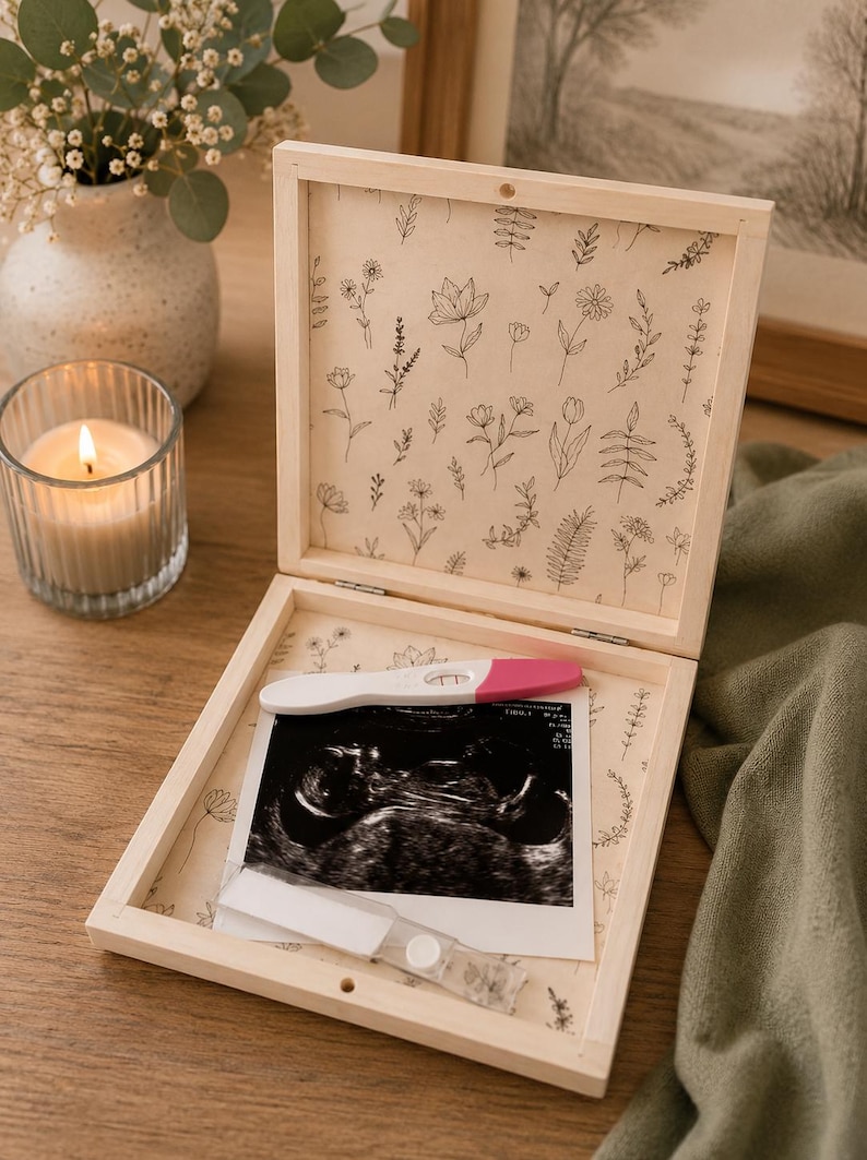 Peut inclure: Coffret souvenir en bois clair, orn&eacute; d'un motif floral &agrave; l'int&eacute;rieur. Il contient un test de grossesse et une &eacute;chographie. Une bougie allum&eacute;e et un vase de fleurs sont en arri&egrave;re-plan.