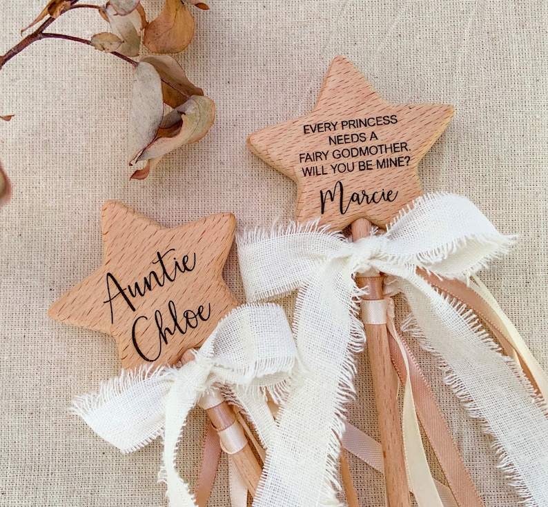 Puede incluir: Dos varitas de madera en forma de estrella con veta clara. Una varita est&aacute; grabada con "Auntie Chloe" y la otra con "EVERY PRINCESS NEEDS A FAIRY GODMOTHER. WILL YOU BE MINE?" y "Marcie". Ambas tienen lazos de cinta color crema deshilachados.