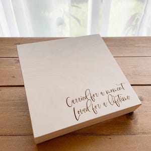 Könnte beinhalten: Eine hellfarbene Holzkiste mit dem eingravierten Text "Carried for a moment, Loved for a lifetime." Die Kiste ist rechteckig und steht auf einer Holzoberfläche, mit einem Fenster und transparenten Vorhängen im Hintergrund.