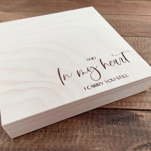 Könnte beinhalten: Eine hellfarbene Holzkiste mit Deckel. Der Deckel ist mit den Worten "In my heart, I carry you still" in einer Schriftart graviert. Die Kiste steht auf einer Holzoberfläche.