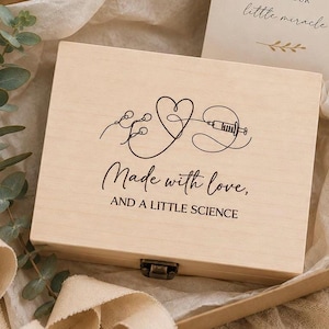 Op de afbeelding: Een rechthoekige, lichtgekleurde houten doos met een metalen sluiting, voorzien van een zwarte lijntekening van een hart verbonden met een injectiespuit en de tekst "Made with love, AND A LITTLE SCIENCE" in een combinatie van script- en schreefloze lettertypes.