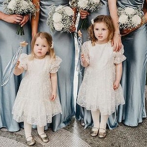Peut inclure: Deux jeunes filles portant des robes en dentelle blanche &agrave; manches bouffantes et des chaussures dor&eacute;es. Elles tiennent des baguettes avec des &eacute;toiles au sommet. Les filles sont debout devant un groupe de femmes portant de longues robes en satin bleu.