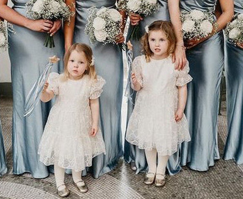 Peut inclure: Deux jeunes filles portant des robes en dentelle blanche &agrave; manches bouffantes et des chaussures dor&eacute;es. Elles tiennent des baguettes avec des &eacute;toiles au sommet. Les filles sont debout devant un groupe de femmes portant de longues robes en satin bleu.