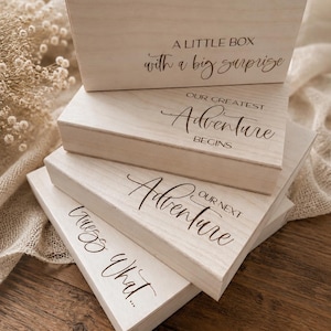 Könnte beinhalten: Ein Stapel aus vier rechteckigen, hellen Holzschachteln mit eingraviertem schwarzem Text. Die Schachteln tragen die Aufschriften A LITTLE BOX with a big surprise, OUR GREATEST Adventure BEGINS, OUR NEXT Adventure und Guess What... in verschiedenen Schriftarten.