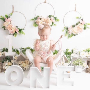 Peut inclure: Un panneau en bois blanc 'UN' avec une petite fille assise sur un tabouret en bois blanc. Le b&eacute;b&eacute; porte une robe blanche avec des fleurs roses et un chapeau rose. Il y a deux couronnes de fleurs accroch&eacute;es au mur derri&egrave;re le b&eacute;b&eacute;.
