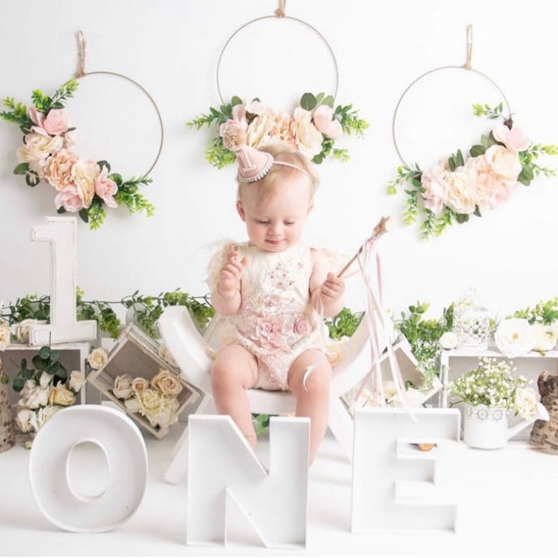 Peut inclure: Un panneau en bois blanc 'UN' avec une petite fille assise sur un tabouret en bois blanc. Le b&eacute;b&eacute; porte une robe blanche avec des fleurs roses et un chapeau rose. Il y a deux couronnes de fleurs accroch&eacute;es au mur derri&egrave;re le b&eacute;b&eacute;.
