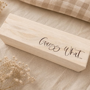 Könnte beinhalten: Eine rechteckige, helle Holzschachtel mit glatter Oberfläche. Auf der Oberseite ist der Text "Guess What..." in einer dunklen Schreibschrift eingraviert. Die Schachtel hat ein schlichtes Design mit sichtbarer, natürlicher Holzmaserung.