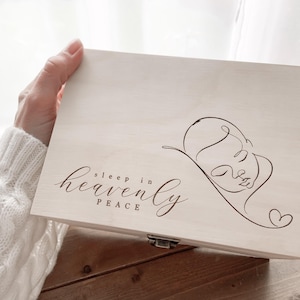 Könnte beinhalten: Eine hellfarbene Holzkiste mit Scharnierdeckel. Die Kiste zeigt eine Strichzeichnung eines schlafenden Babys und die Worte "sleep in heaven peace" in eleganter Schrift. Ein kleines Herz ist in der Nähe des Babys gezeichnet.