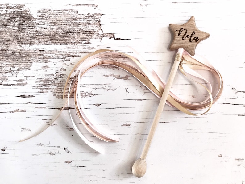 Peut inclure: Une baguette en bois en forme d'&eacute;toile avec un ruban rose et or attach&eacute;. L'&eacute;toile est grav&eacute;e du nom "Nola".