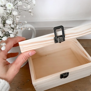 May include: A small, unfinished wooden box with a hinged lid, open to reveal the interior. The box has a metal clasp and hinges. The lid is being held open by a hand. The box is sitting on a wooden surface.