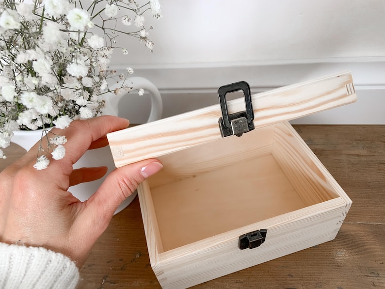 May include: A small, unfinished wooden box with a hinged lid, open to reveal the interior. The box has a metal clasp and hinges. The lid is being held open by a hand. The box is sitting on a wooden surface.