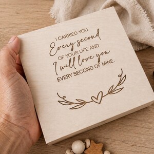 Könnte beinhalten: Eine quadratische, helle Holzplatte mit dem lasergravierten Text: I CARRIED YOU Every second OF YOUR LIFE AND I will love you EVERY SECOND OF MINE. Unter dem Text befindet sich eine dekorative Strichzeichnung eines Herzens zwischen zwei Blattzweigen.
