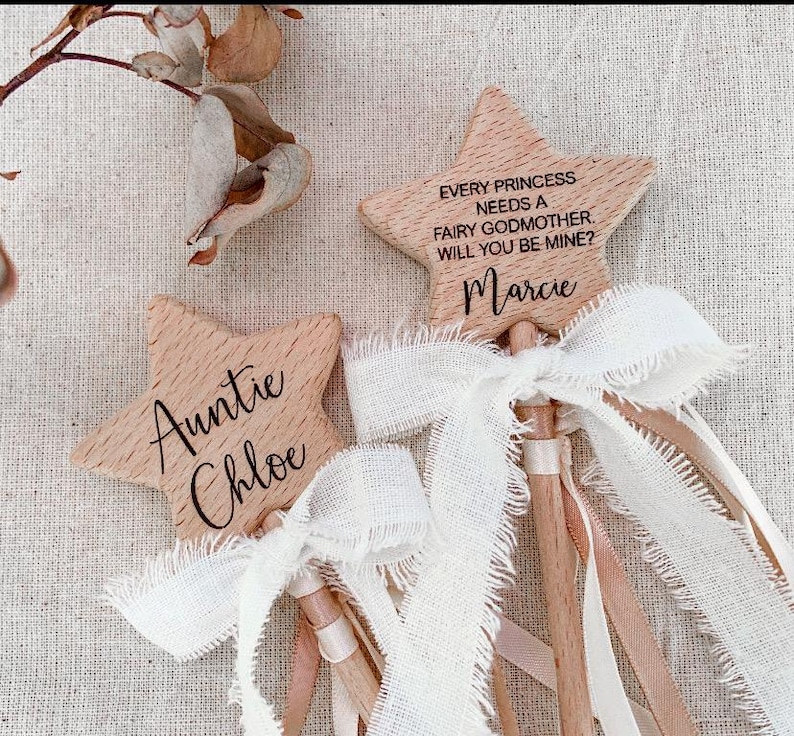 Puede incluir: Dos varitas de madera en forma de estrella con veta clara. Una varita est&aacute; grabada con "Auntie Chloe" y la otra con "EVERY PRINCESS NEEDS A FAIRY GODMOTHER. WILL YOU BE MINE? Marcie". Ambas tienen lazos de cinta blanca deshilachada.