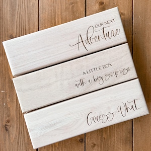 Könnte beinhalten: Drei rechteckige, weiß getünchte Holzkisten mit dunkelbraunen Kanten. Die Kisten tragen die Aufschriften "OUR NEXT Adventure", "A LITTLE BOX with a big surprise" und "Guess What..." in Schreibschrift.