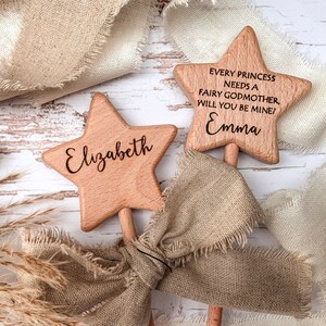Puede incluir: Dos varitas de madera de color marr&oacute;n claro en forma de estrella con palos. Una varita est&aacute; grabada con el nombre Elizabeth. La otra varita tiene grabado el texto Every princess needs a fairy godmother, will you be mine? y el nombre Emma en negro.